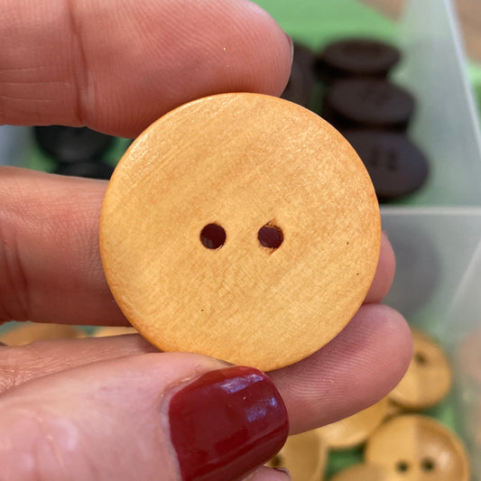 Hand holding a round wooden button with two holes, with a container of buttons in the background.