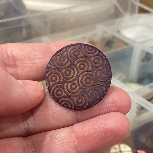 Hand holding a decorative button with a pattern, against a background of a container with various buttons.