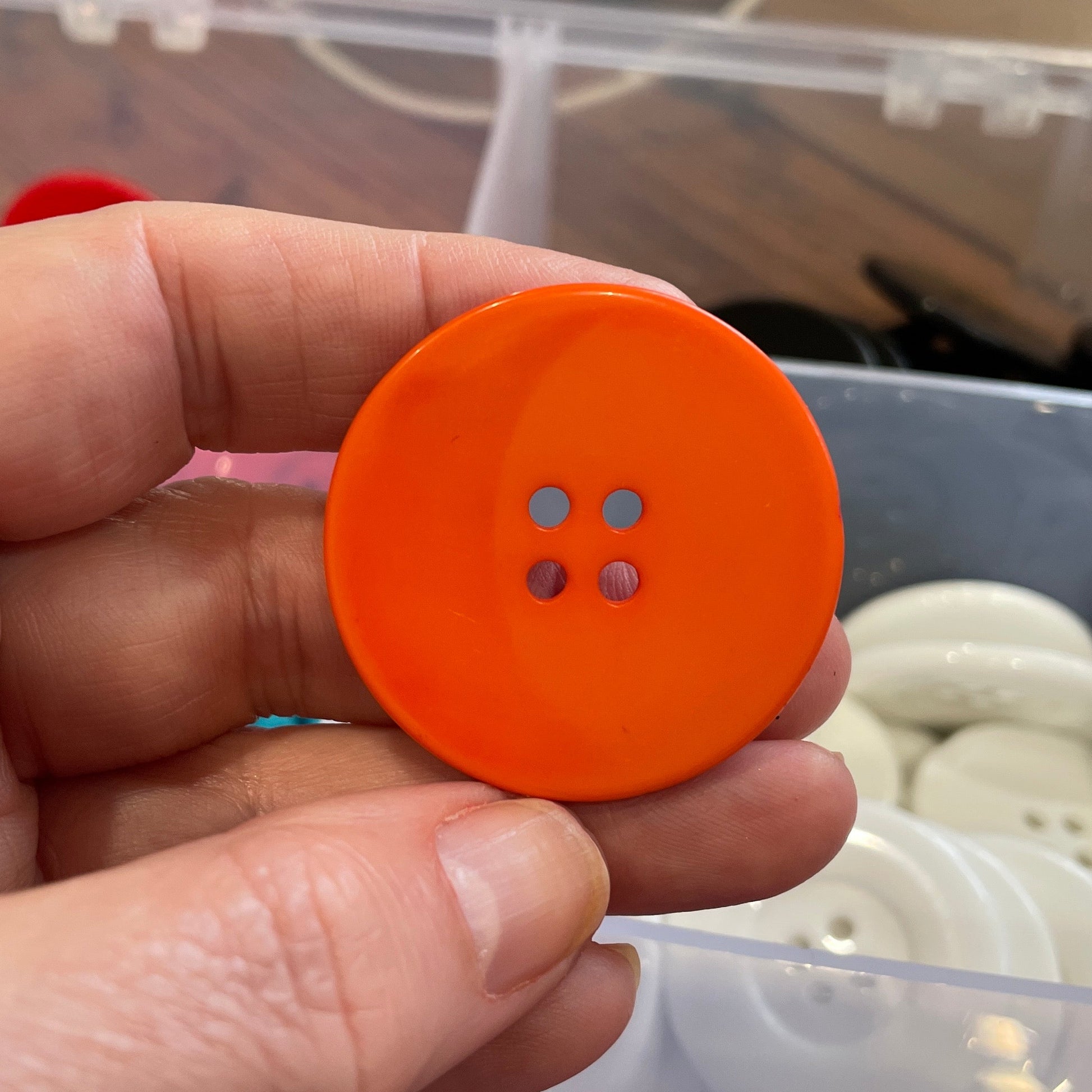 Hand holding an orange button with two holes in front of a container of buttons.