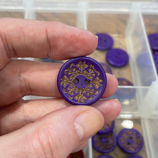 Hand holding a purple button with gold design in front of a container of buttons.