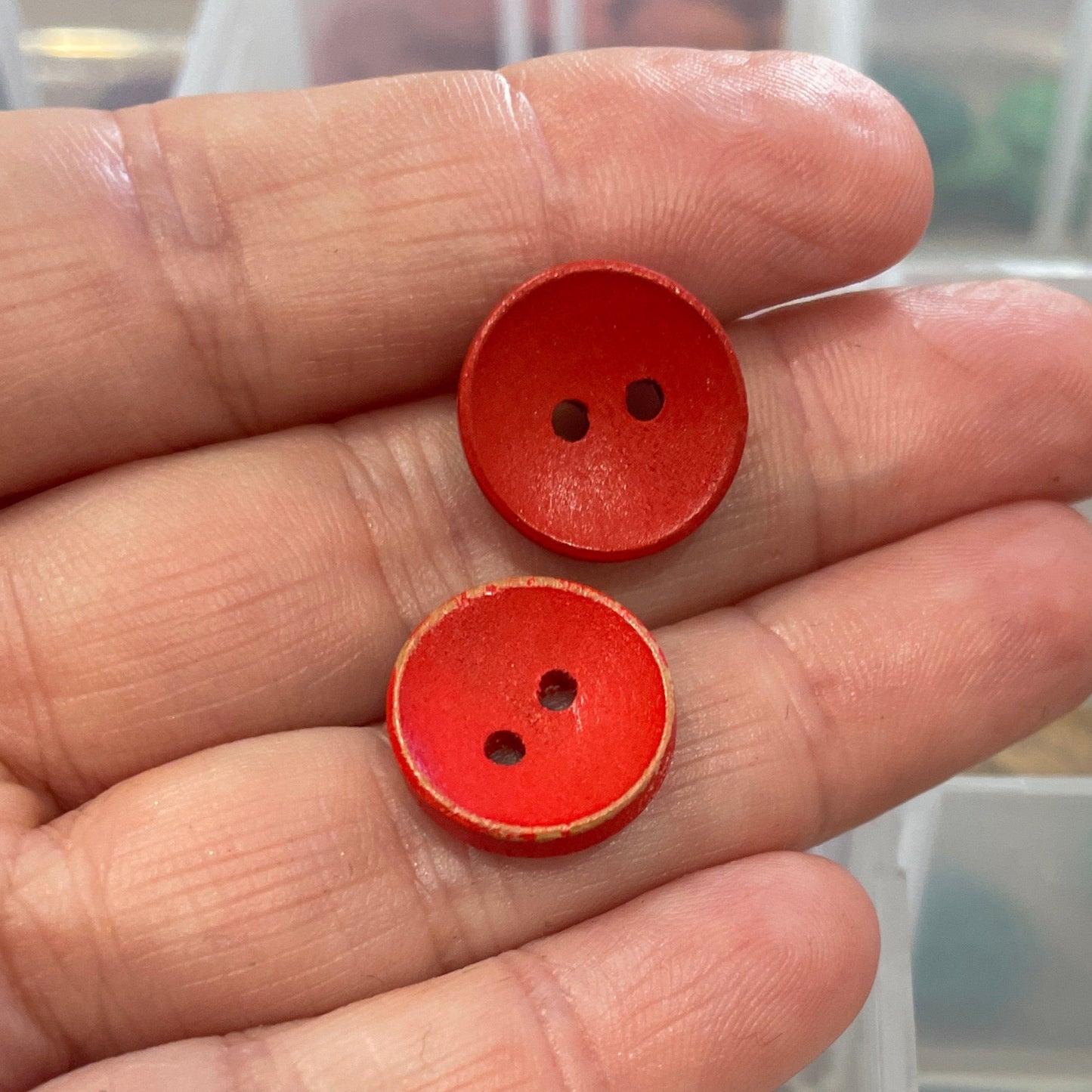 Hand holding two red buttons with a clear container of assorted buttons in the background.