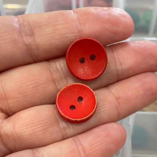 Hand holding two red buttons with a clear container of assorted buttons in the background.