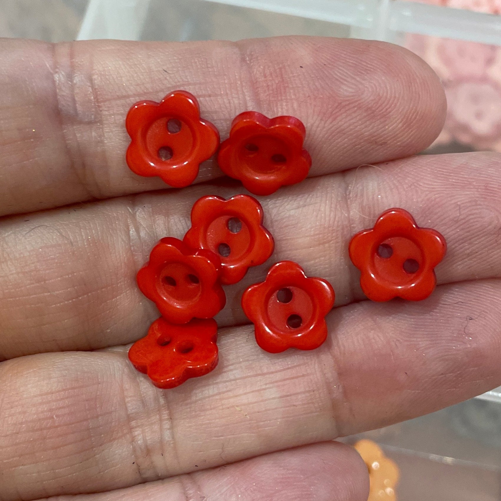 Hand holding red flower-shaped buttons with a container of similar buttons in the background.