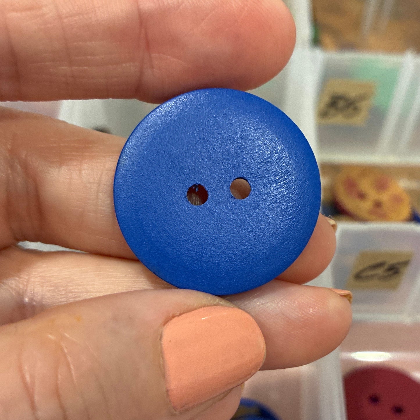 Hand holding a blue button with a background of button storage containers.