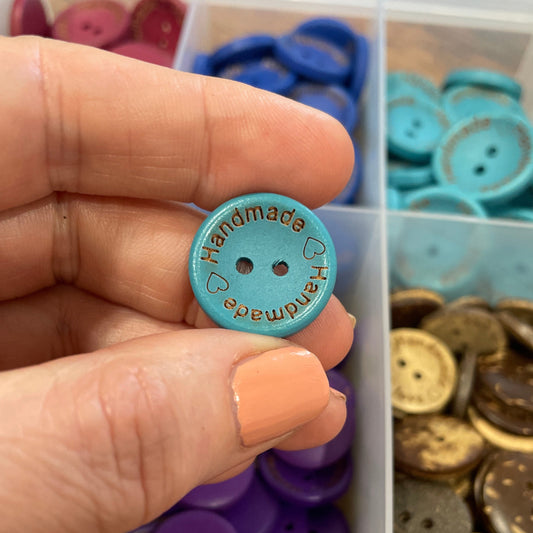 Hand holding a blue button with 'Handmade' text, against a background of assorted buttons.