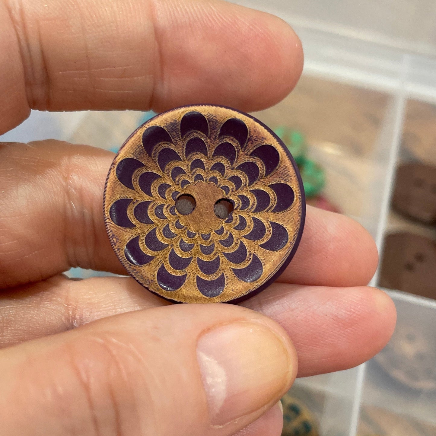 Hand holding a wooden button with intricate design, with a box of similar buttons in the background.