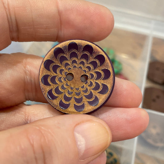 Hand holding a wooden button with intricate design, with a box of similar buttons in the background.