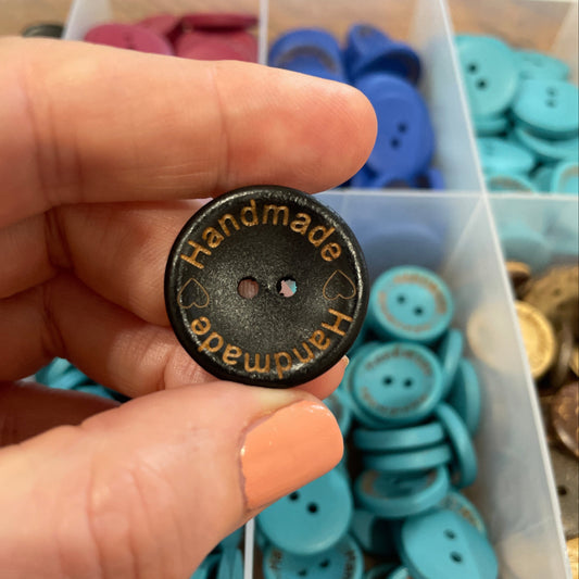 Hand holding a button with 'Handmade' text in front of a container of assorted buttons.