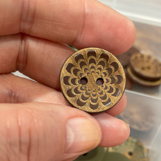 Hand holding a decorative button with a detailed design, set against a background of similar buttons in a container.