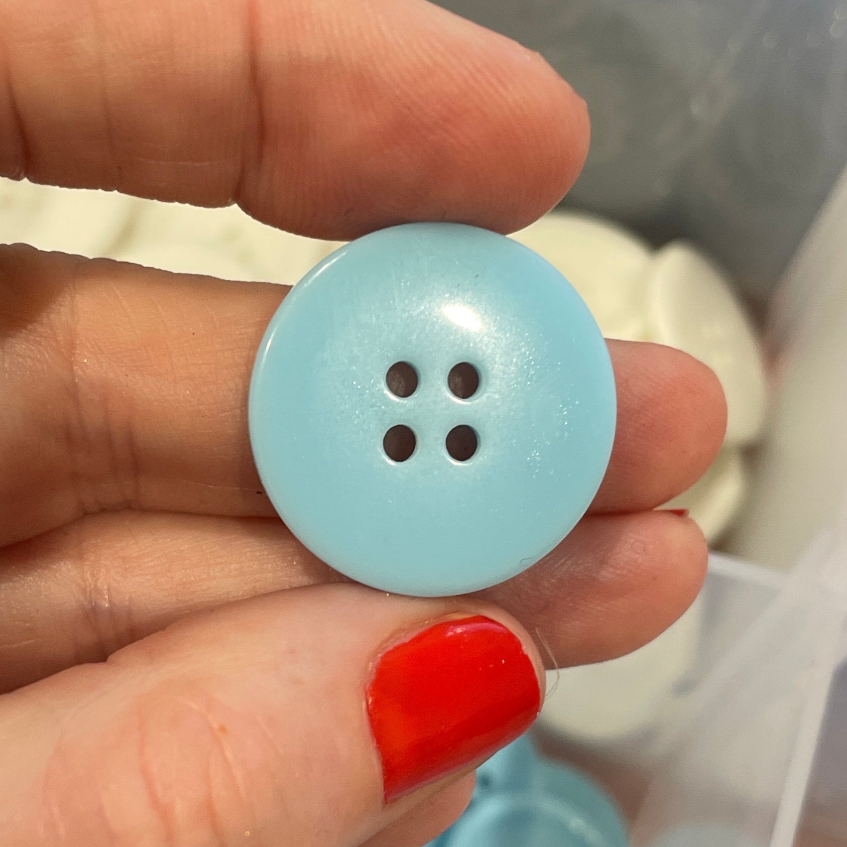 Hand holding a light blue button with a clear container of buttons in the background