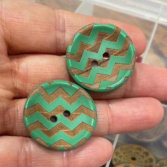 Hand holding two decorative buttons with a patterned container in the background