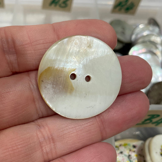 Hand holding a round, white button with two holes against a blurred background of more buttons.