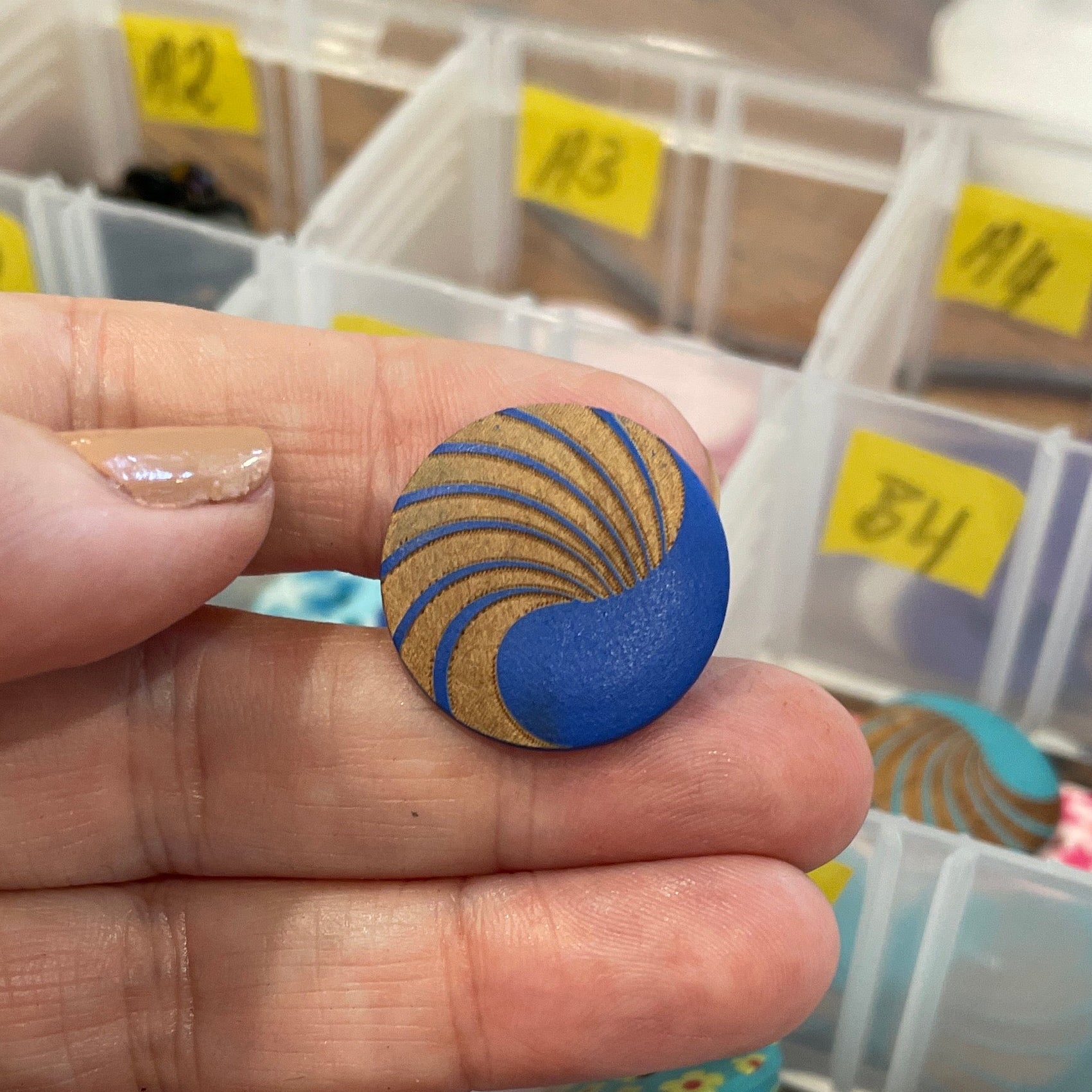 Hand holding a blue button with a yellow pattern against a background of colorful buttons in a container.