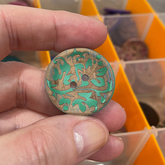 Hand holding a decorative button with intricate design in front of a button collection organizer.