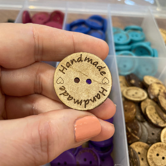 Handmade wooden button held by a hand with a container of buttons in the background.