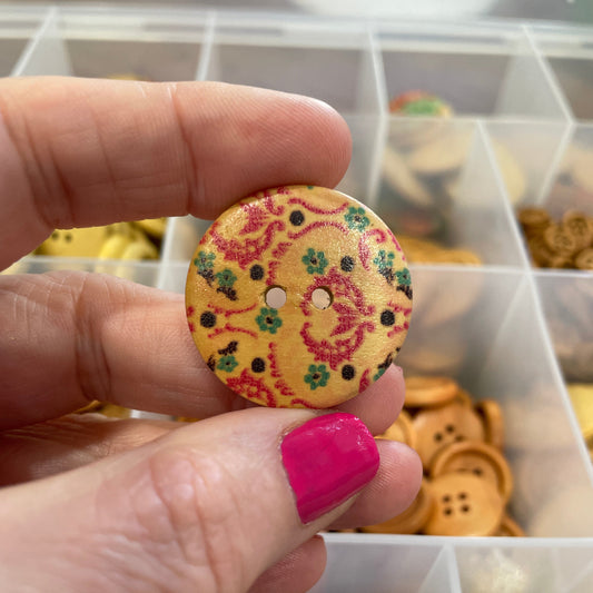 Hand holding a decorative button with a colorful pattern in front of a container of buttons.
