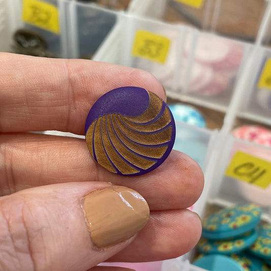 Hand holding a purple and gold button with a container of buttons in the background