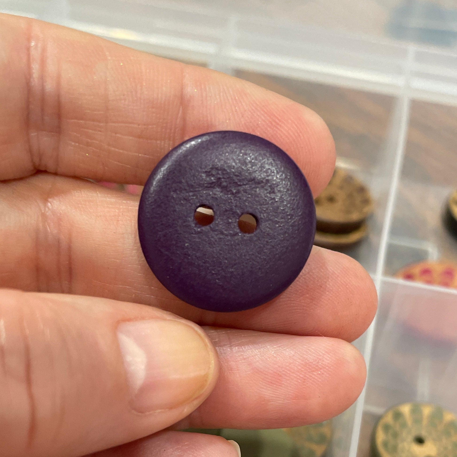 Hand holding a purple button with a container of various buttons in the background