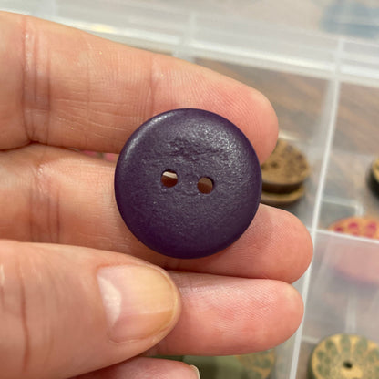 Hand holding a purple button with a container of various buttons in the background