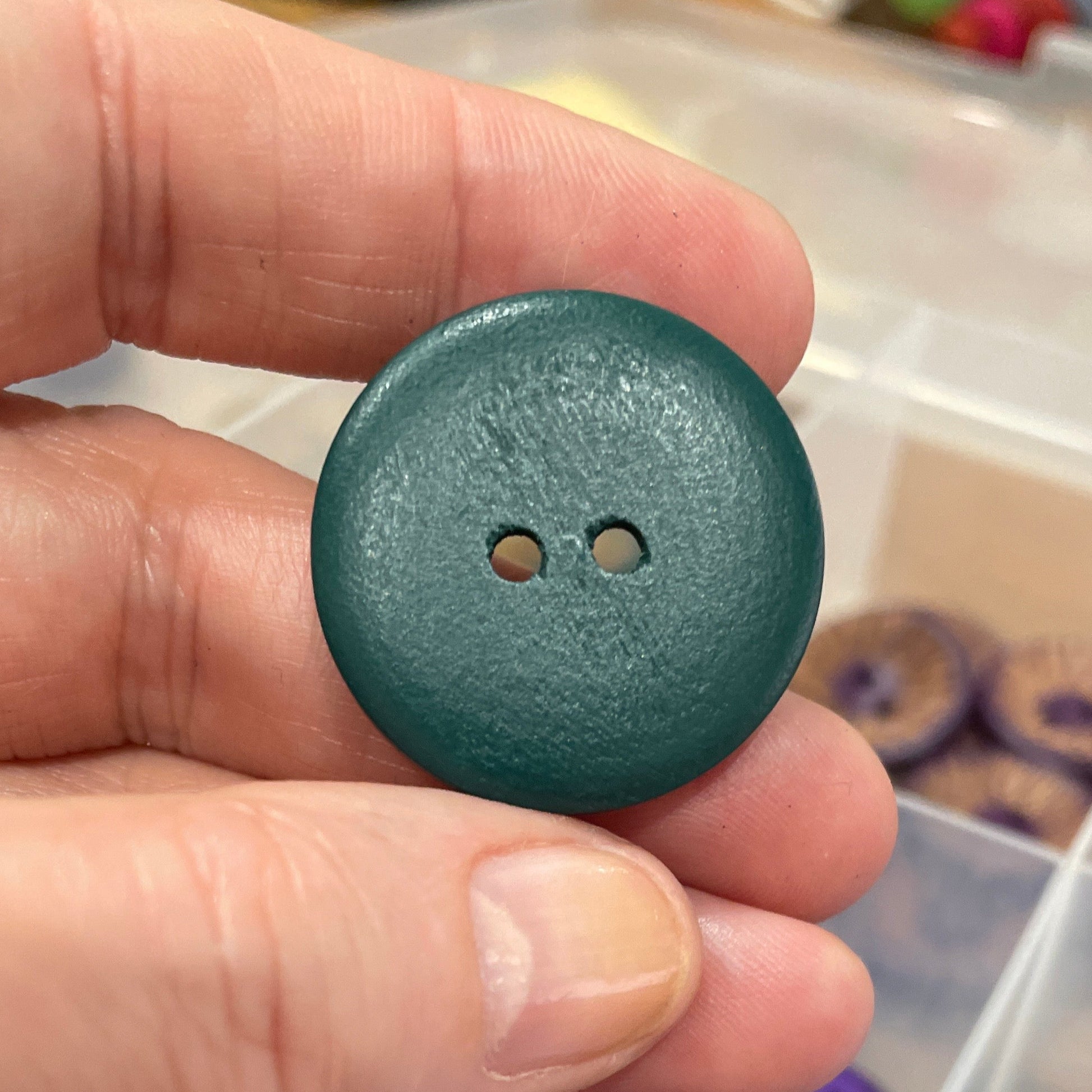 Hand holding a teal button with a container of buttons in the background