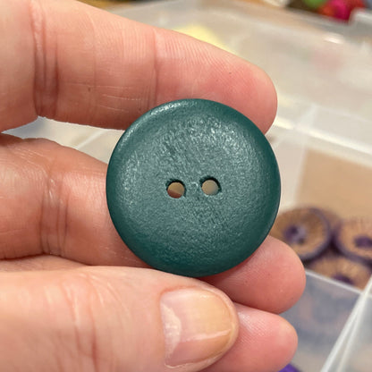 Hand holding a teal button with a container of buttons in the background