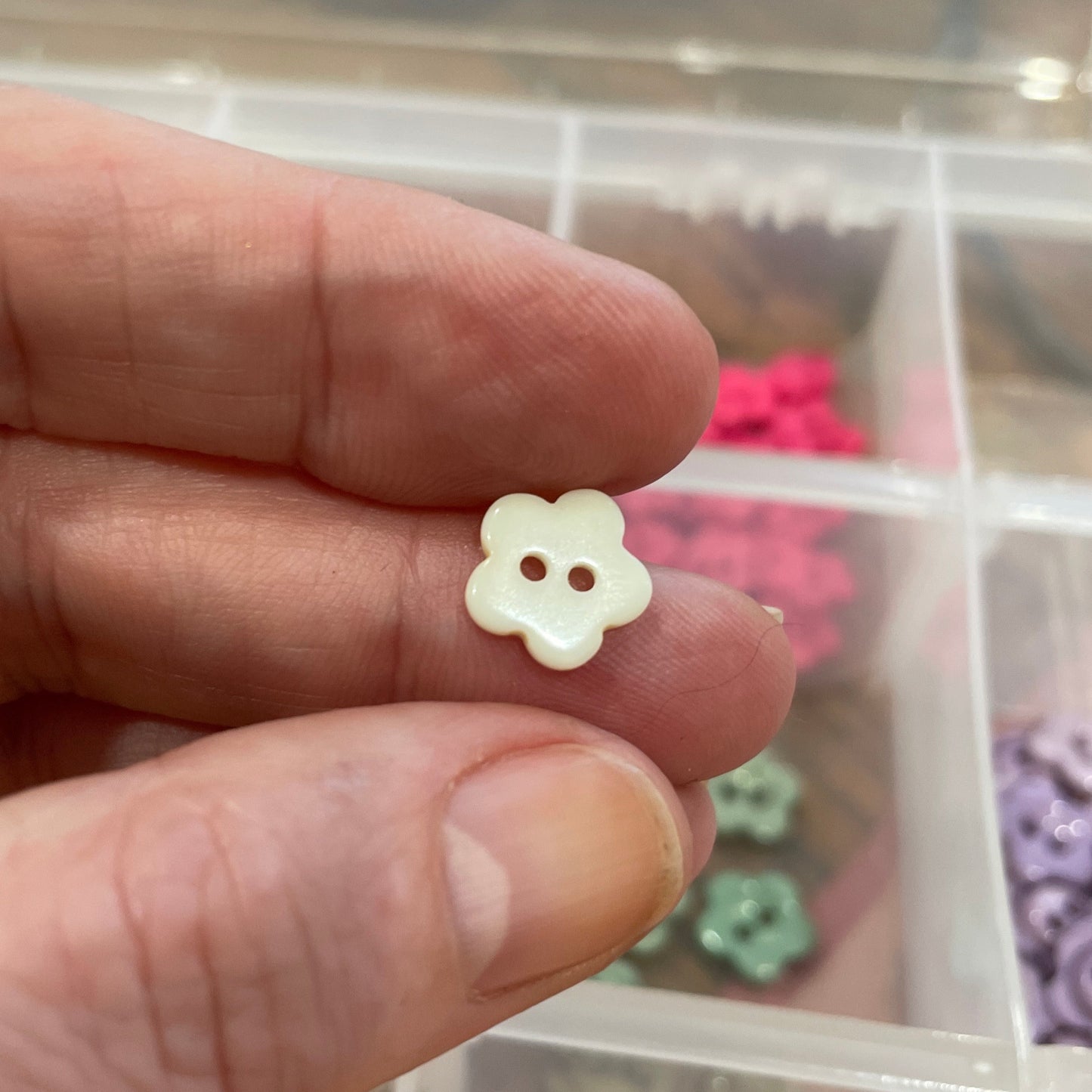 Hand holding a small white flower-shaped button with a container of colorful buttons in the background.