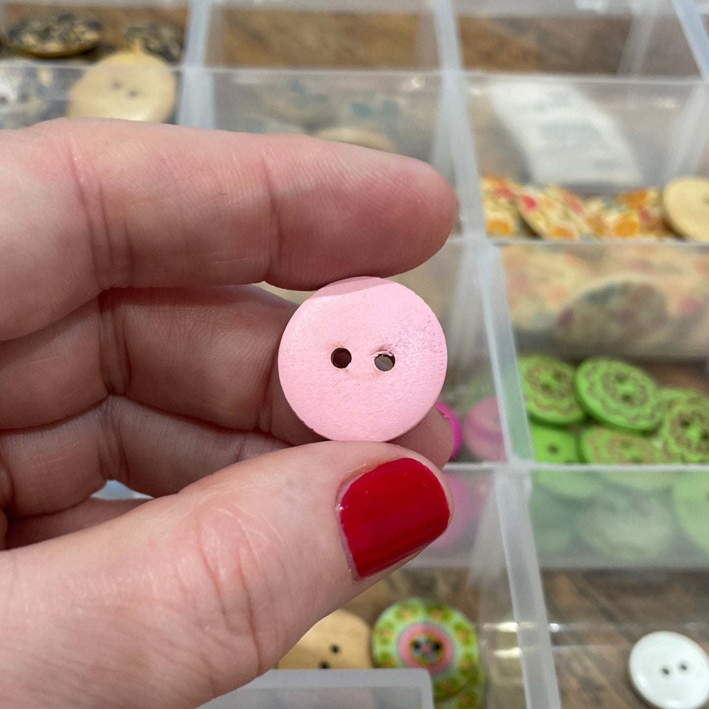 Hand holding a pink button with a clear container of buttons in the background
