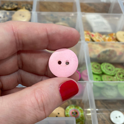 Hand holding a pink button with a clear container of buttons in the background