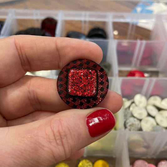 Hand holding a red glittery button with a container of colorful buttons in the background.