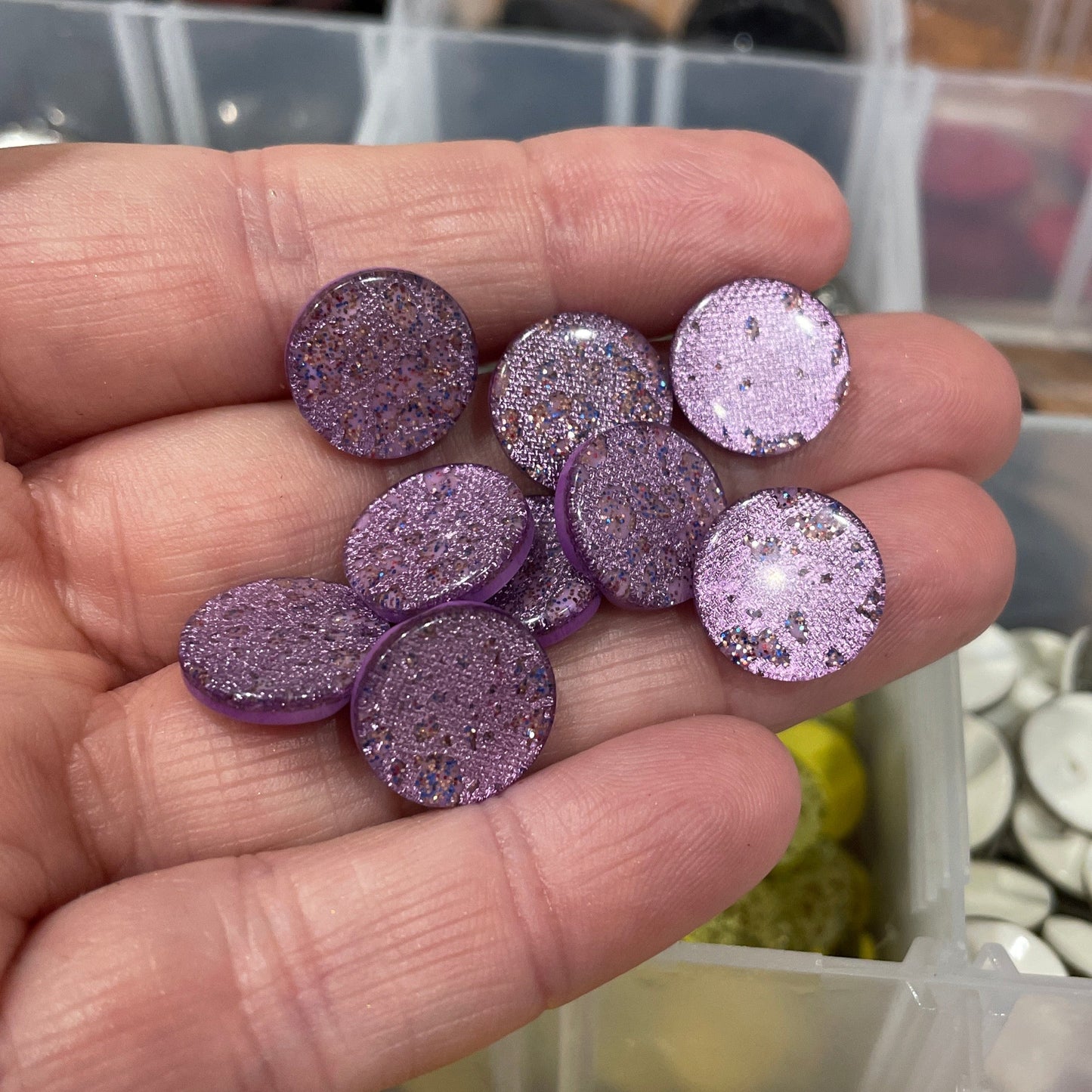 Hand holding purple glittery stones with a background of various colored stones in containers.