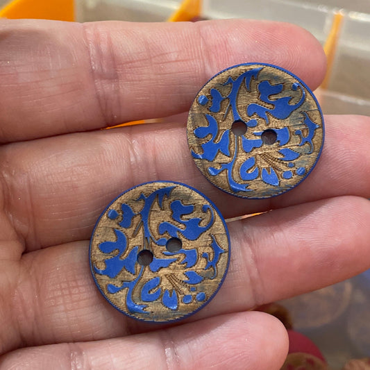 Hand holding two decorative buttons with a blurred background of a button collection box.