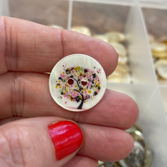 Hand holding a decorative button with a colorful design, surrounded by a container of buttons.