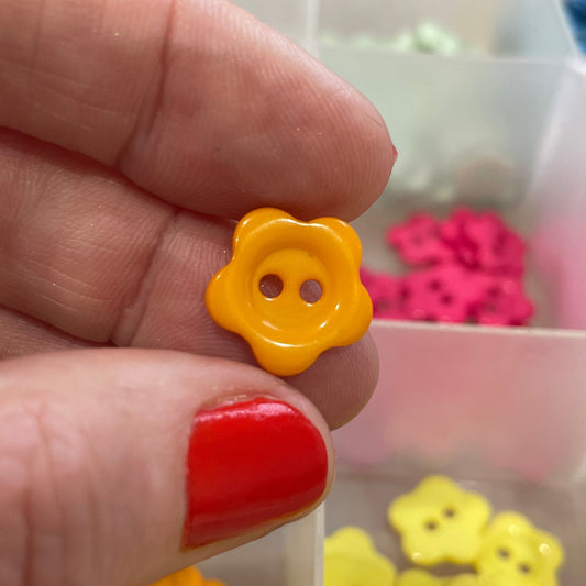 Yellow flower-shaped button held between fingers with a container of similar buttons in the background.
