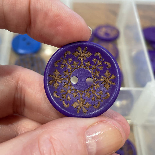 Hand holding a purple button with gold design in front of a container of buttons.