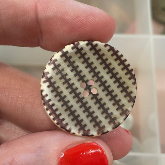 Hand holding a round button with a checkered pattern in front of a container of buttons.