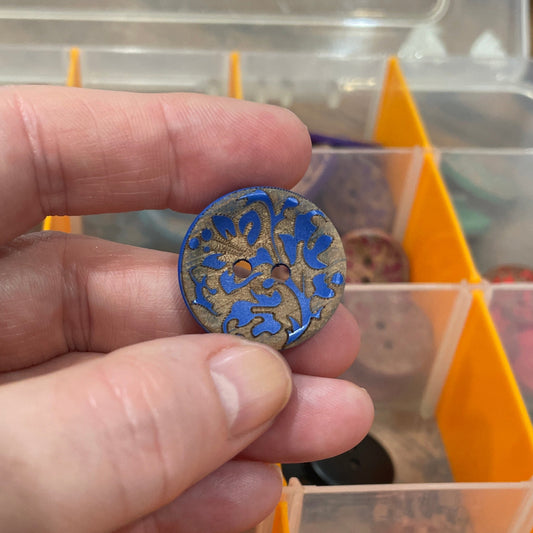 Hand holding a decorative button with a colorful pattern, in front of a container filled with various buttons.