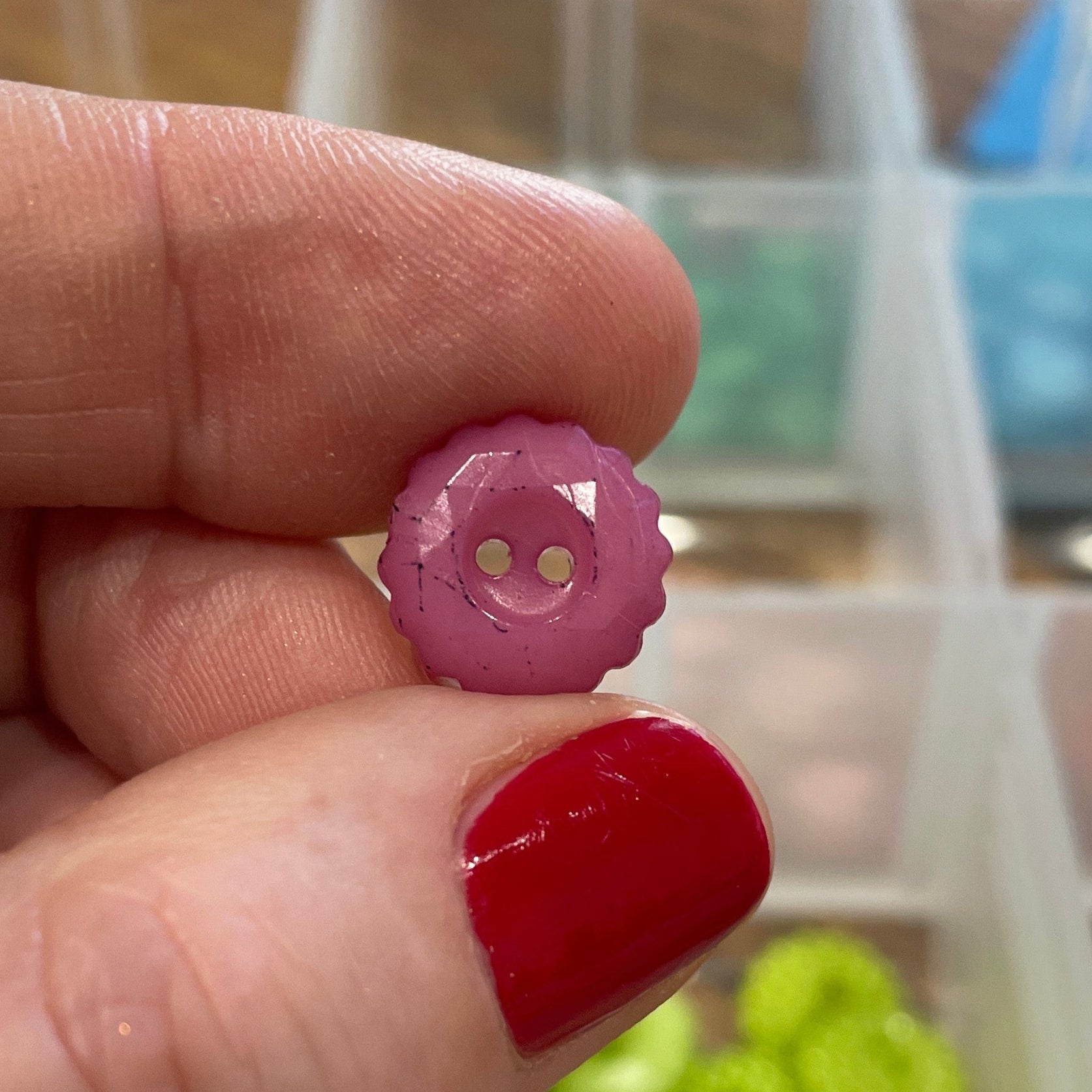 Hand holding a small pink button with a blurred background of a button collection box.