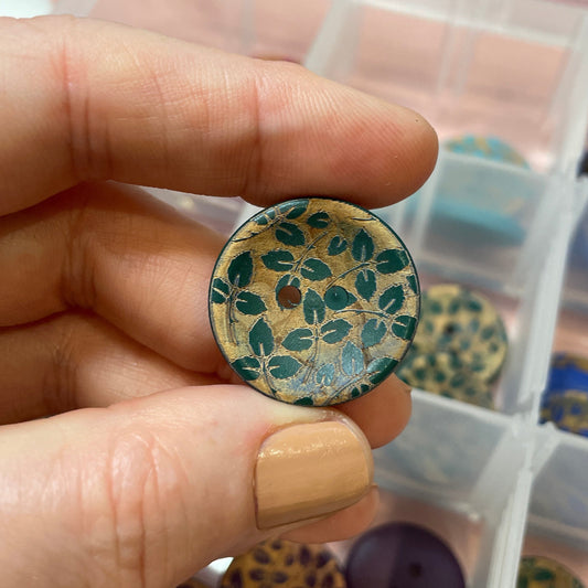 Hand holding a decorative button with a leopard print pattern over a container of similar buttons.
