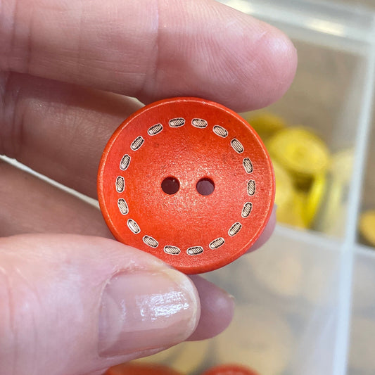 Orange button held between fingers with a container of buttons in the background