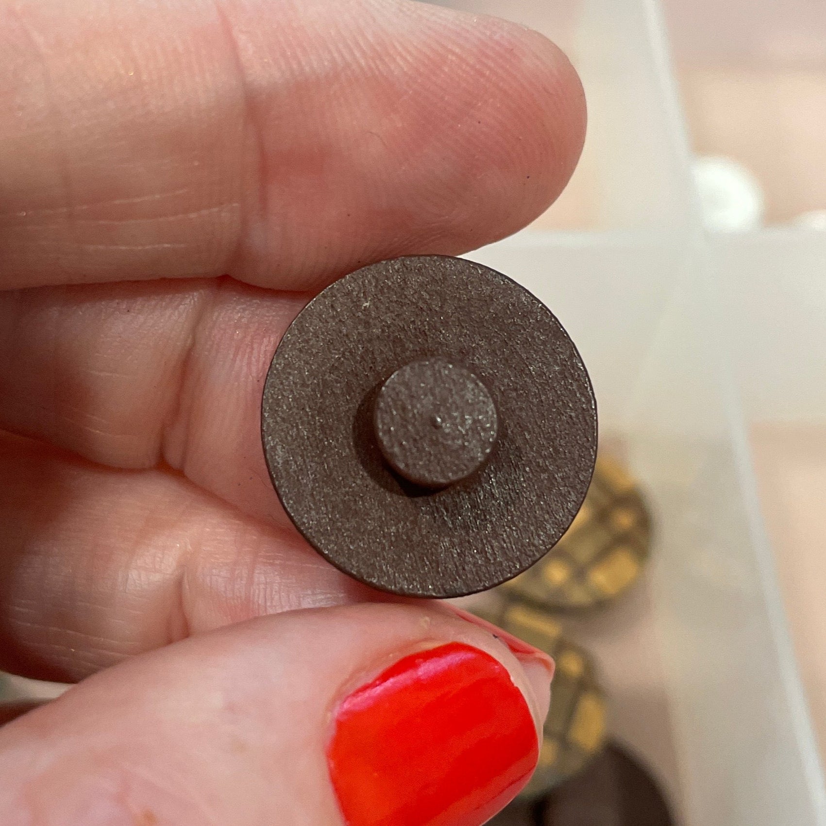 Person holding a small round metal object with a textured surface, against a background of a divided container with various items.