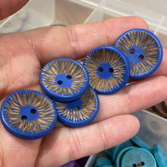 Hand holding blue buttons with intricate designs, with a container of buttons in the background.