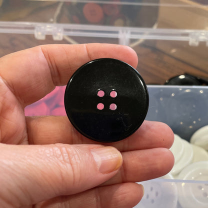 Hand holding a black button in front of a container of buttons.