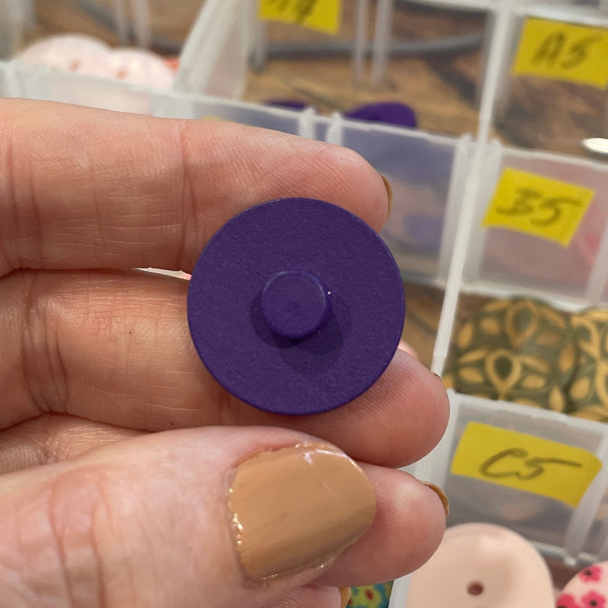 Hand holding a purple button with a background of button containers.
