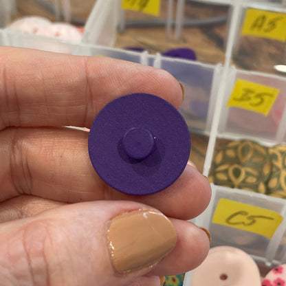 Hand holding a purple button with a background of button containers.
