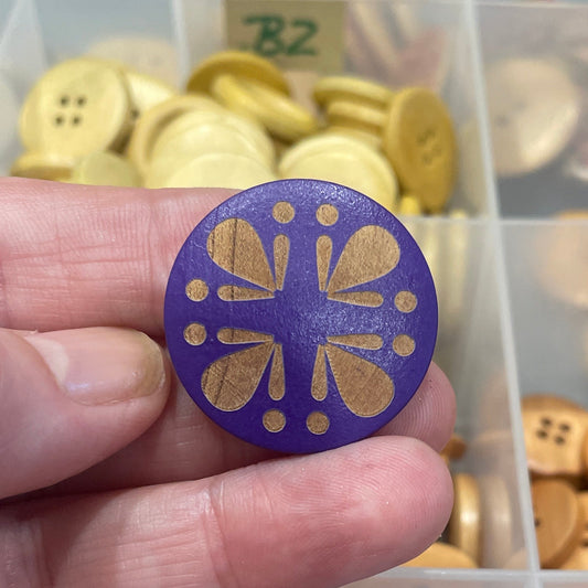 Hand holding a decorative button with a purple and gold design, against a background of various buttons in a container.