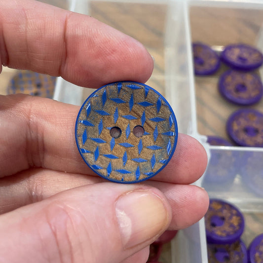 Hand holding a blue button with intricate design, with a container of buttons in the background.