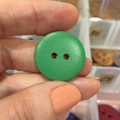 Hand holding a green button with two holes in front of a container of buttons.