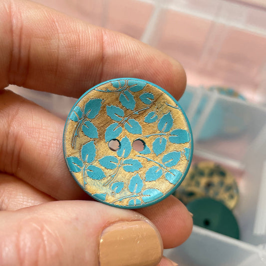 Hand holding a decorative button with intricate design, surrounded by other buttons in a container.