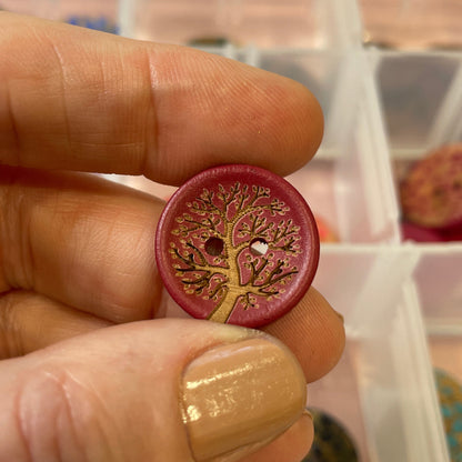 Hand holding a decorative button with a tree of life design in front of a display case with various buttons.