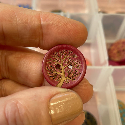 Hand holding a decorative button with a tree of life design in front of a display case with various buttons.